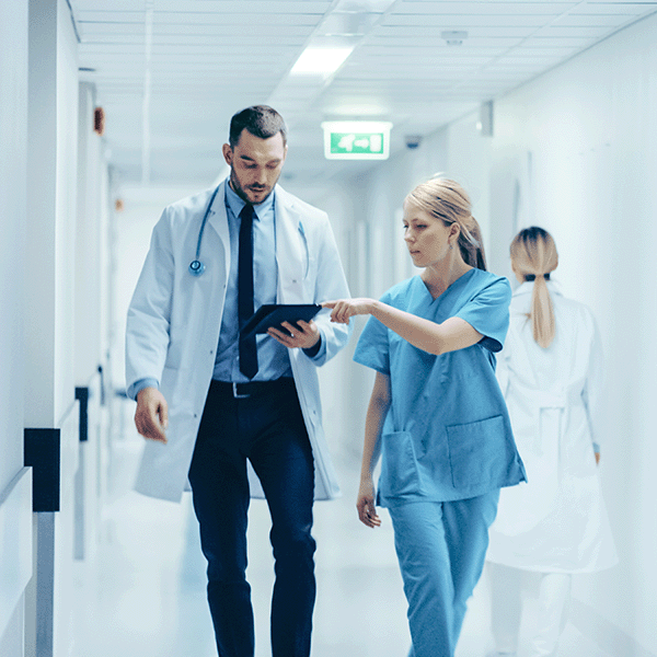 Female nurse in blue scrubs points to a patient chart on the doctor's tablet while they walk down the hospital hallway